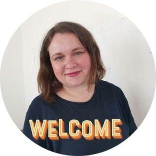 Frau Krone Eine lächelnde Frau mit braunen Haaren trägt ein T-Shirt und steht vor einem hellen Hintergrund.